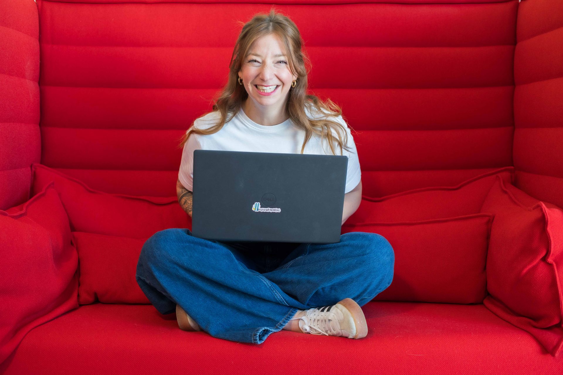 TGIM_2025_ARau-04968 Tanja sitzt lächelnd mit Laptop auf rotem Sofa – Symbolbild für positive Teamkultur, Zusammenarbeit und moderne Führung im Unternehmen.
