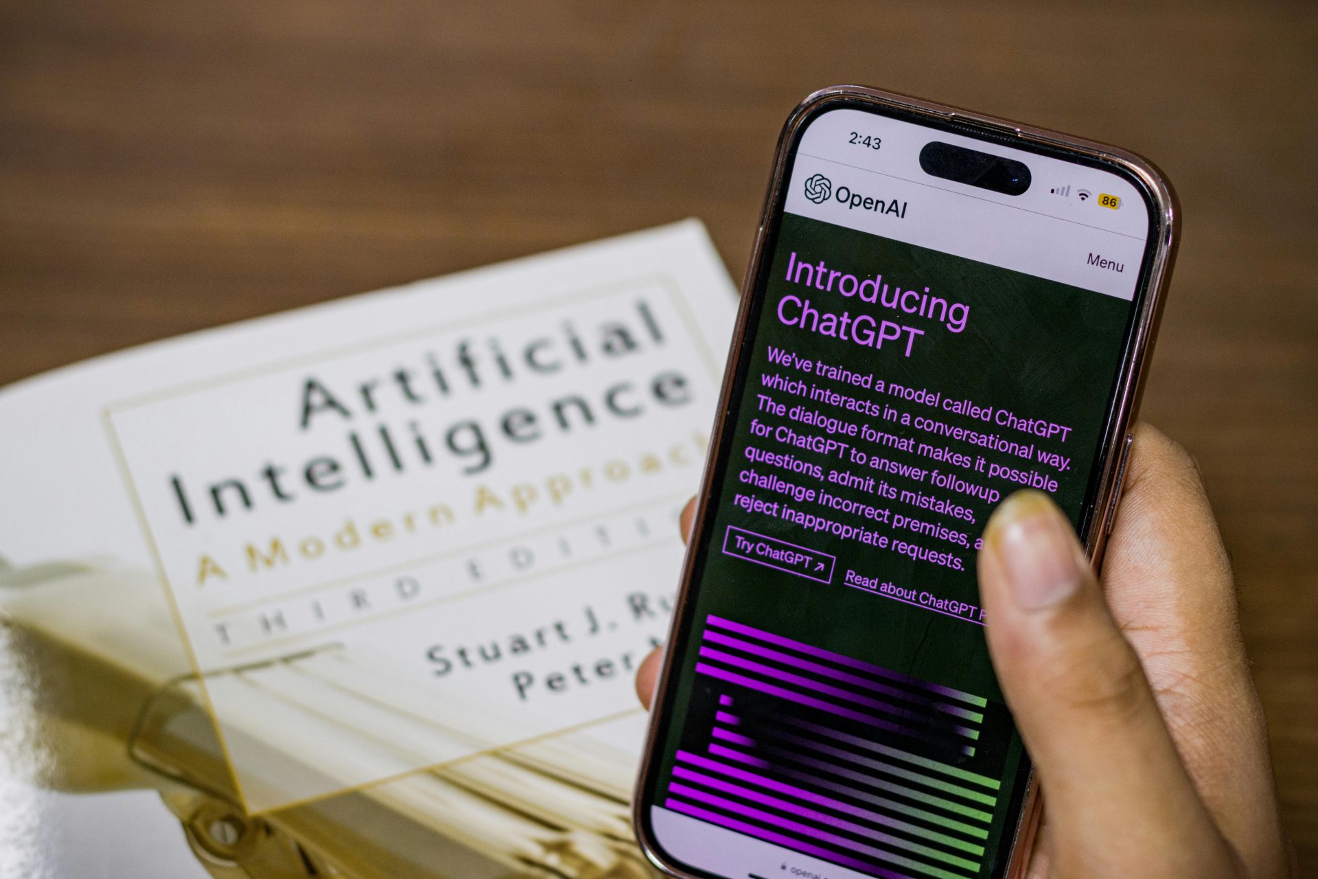 Person hält ein Smartphone mit der OpenAI ChatGPT-Startseite, daneben liegt ein Buch über künstliche Intelligenz (AI)
